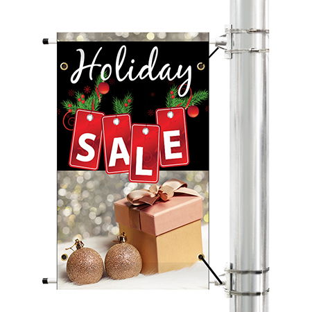 Holiday Sale Pole Banner. It sits to the left of the light pole. Banner is black on top and tan on bottom. The first line of text is in white cursive lettering and reads "Holiday". The next line of text has individual gift tag graphics for each letter of the word "Sale" each letter is white and all caps. Bottom half of the banner has two wooden ornaments to the left, a gold gift box with a pink top in gold gift ribbon sits to the right.
